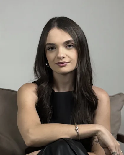 Manoela Grizatti sentada numa poltrona bege, de blusa preta, com os braços apoiados nos joelhos, sobre fundo cinza